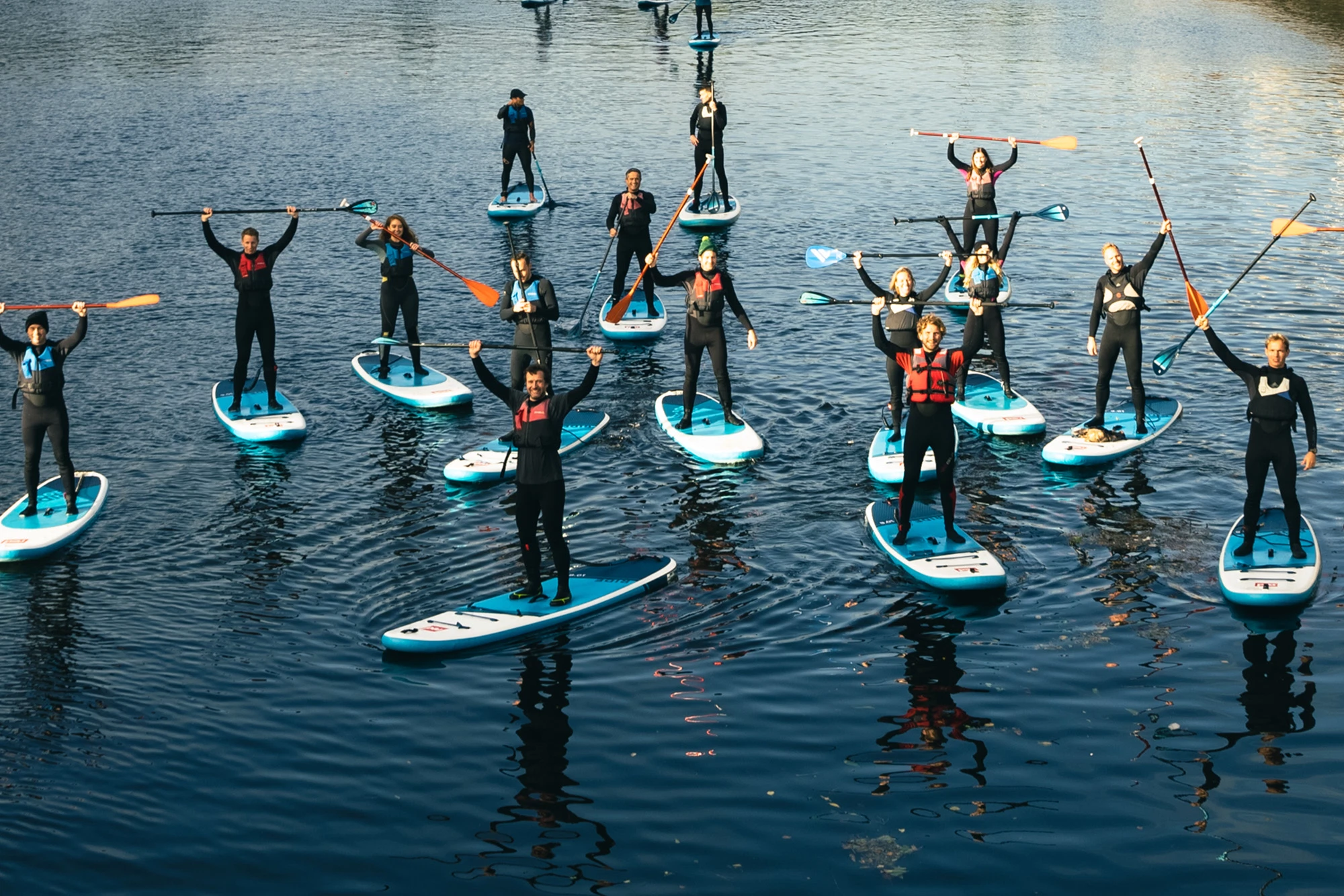 Group paddleboarding on calm water, participants in wetsuits holding paddles high in a celebratory pose.