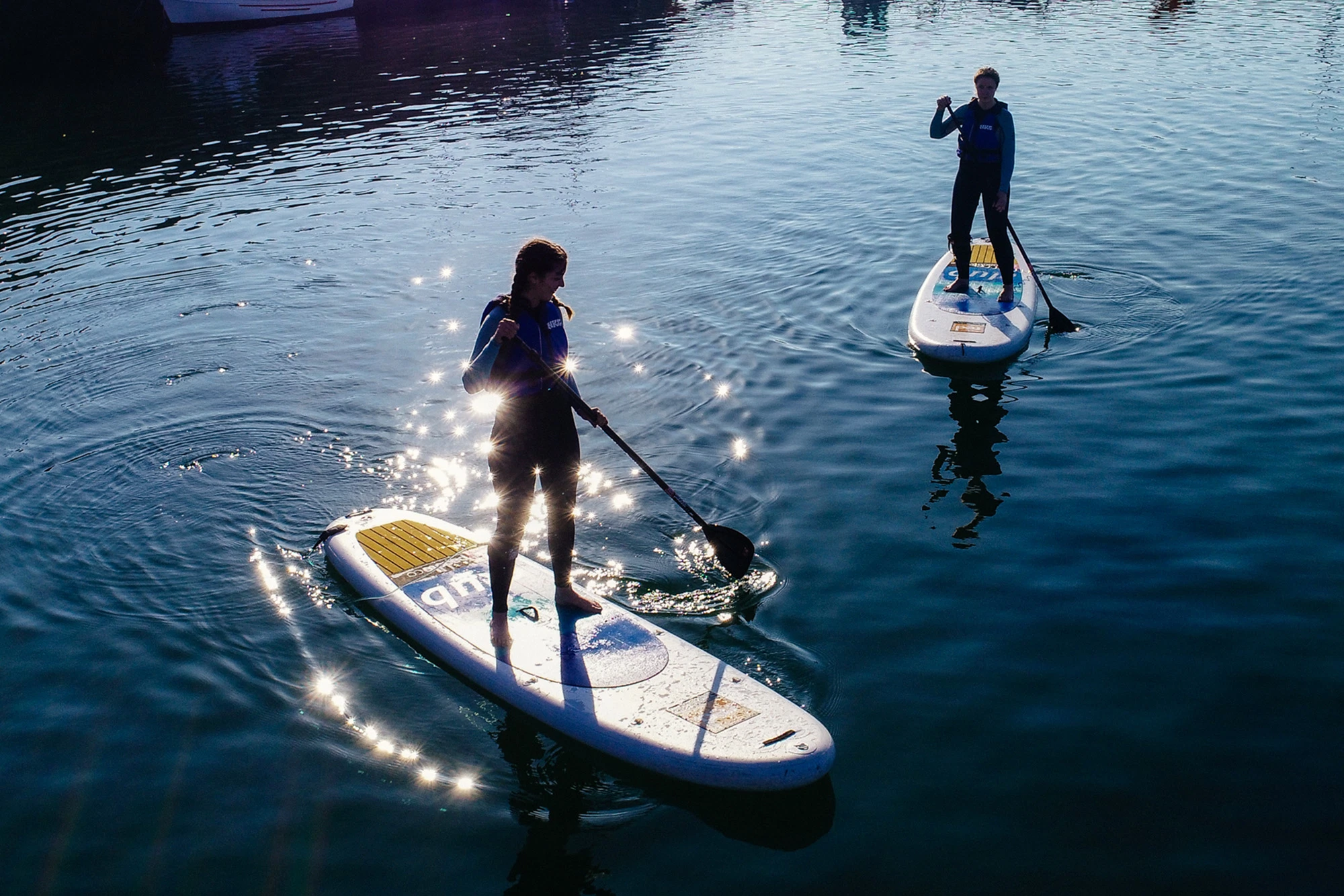 Two people paddleboarding on a sunlit lake, wearing wetsuits, gliding through sparkling water.
