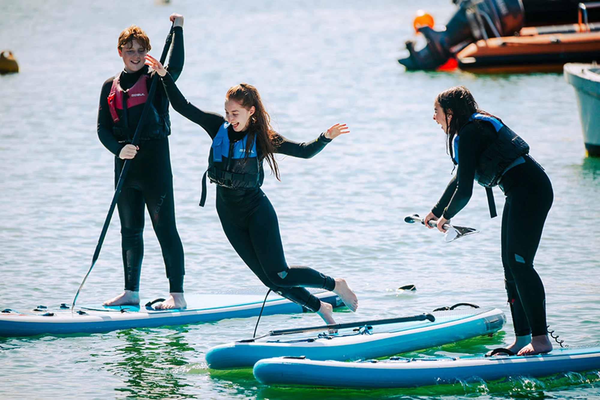 Three people paddleboarding, one jumping into water, all in wetsuits and lifejackets, on a sunny day.
