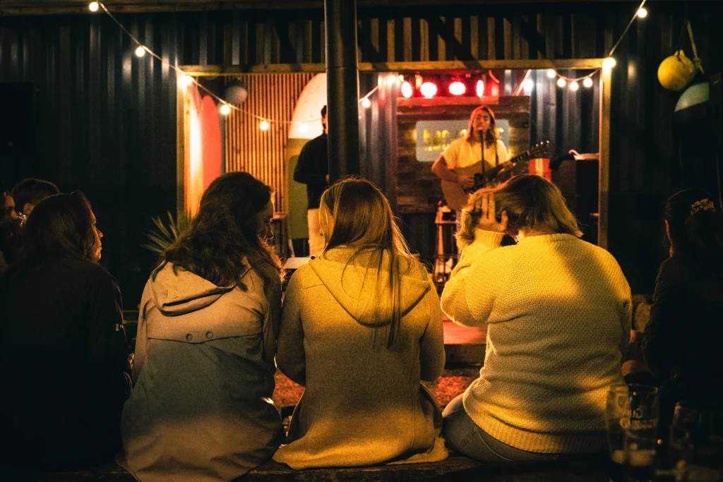 Audience enjoys live music performance at cozy outdoor venue with string lights.