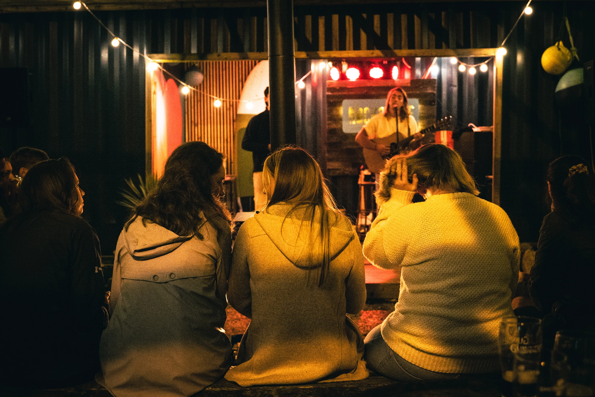 Audience enjoys live music performance at cozy outdoor venue with string lights.