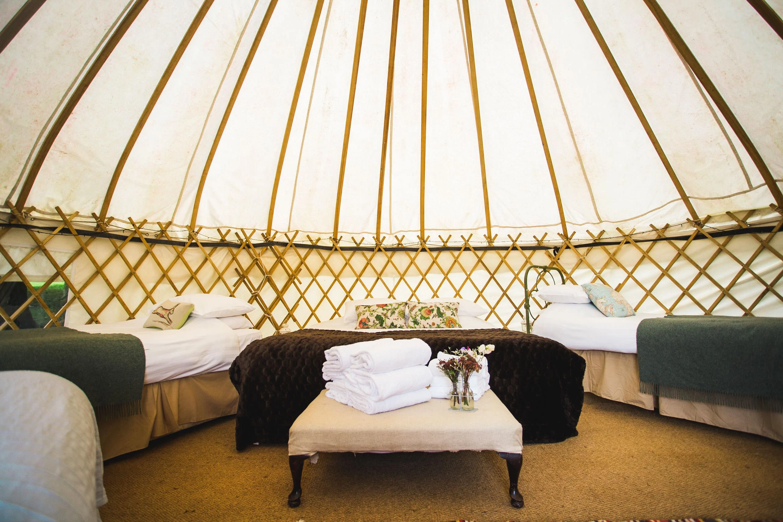Interior of spacious glamping yurt with cosy beds, wooden frame, beige decor, and fresh flowers.