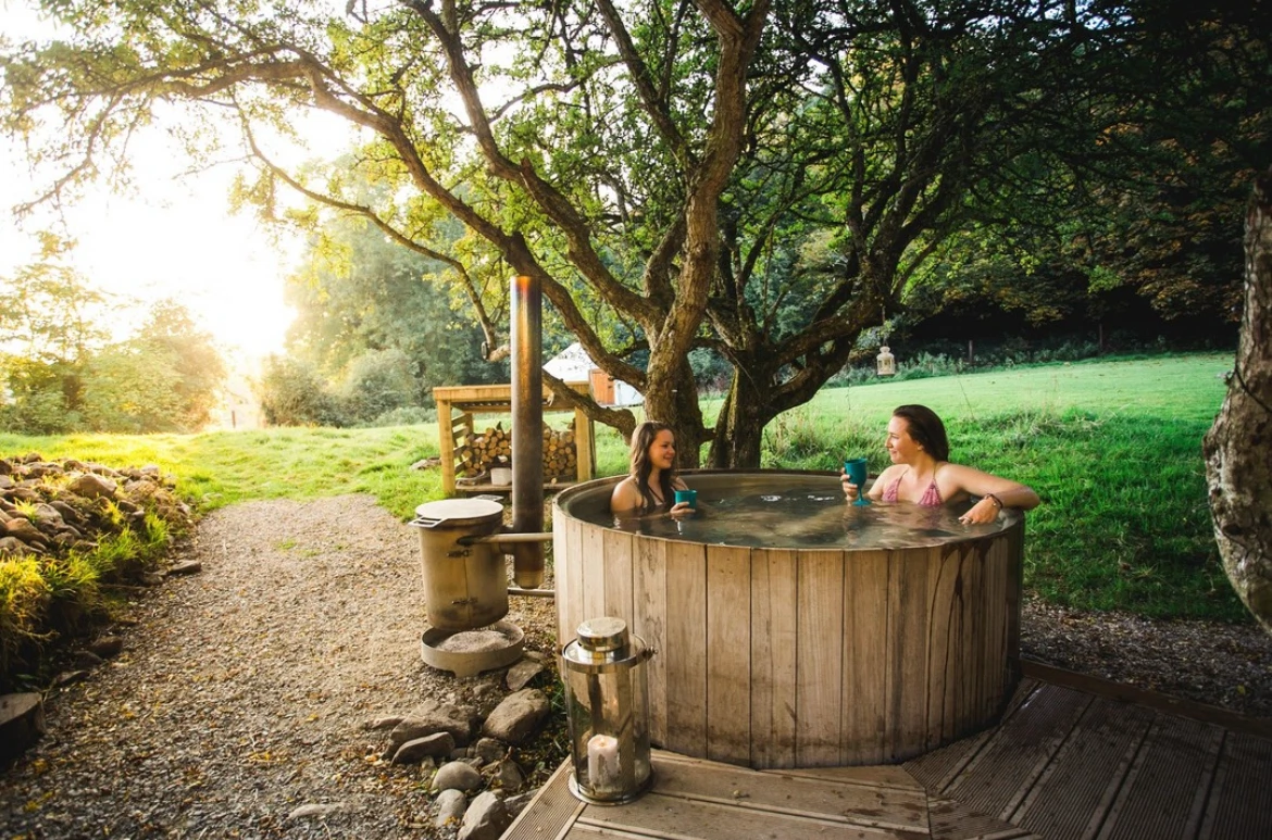 Two women relax in a wooden hot tub outdoors, surrounded by trees and grass under a golden sunset.