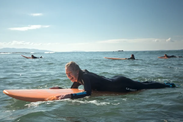 Surfer in wetsuit smiling, lying on orange surfboard in ocean, with others in background. Sunny day, calm sea.