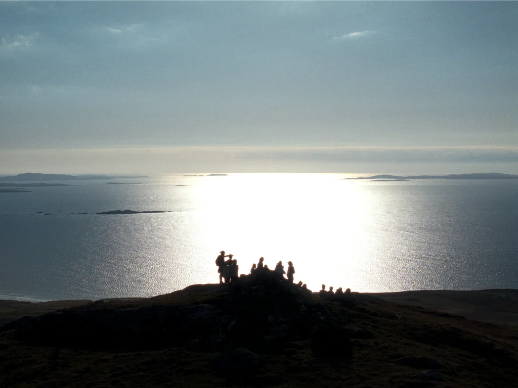 Silhouette of hikers on hill with panoramic coastal sunset view, shimmering sea and distant islands.