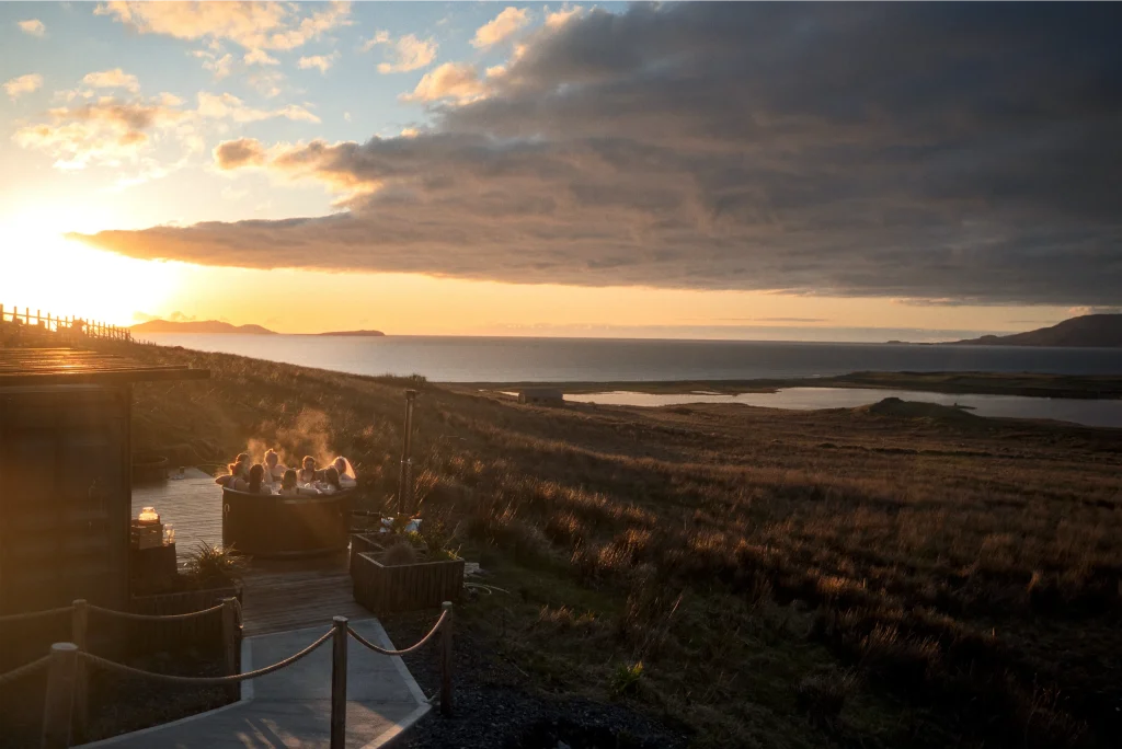 People in a hot tub on a scenic hilltop at sunset, overlooking the ocean and landscape.