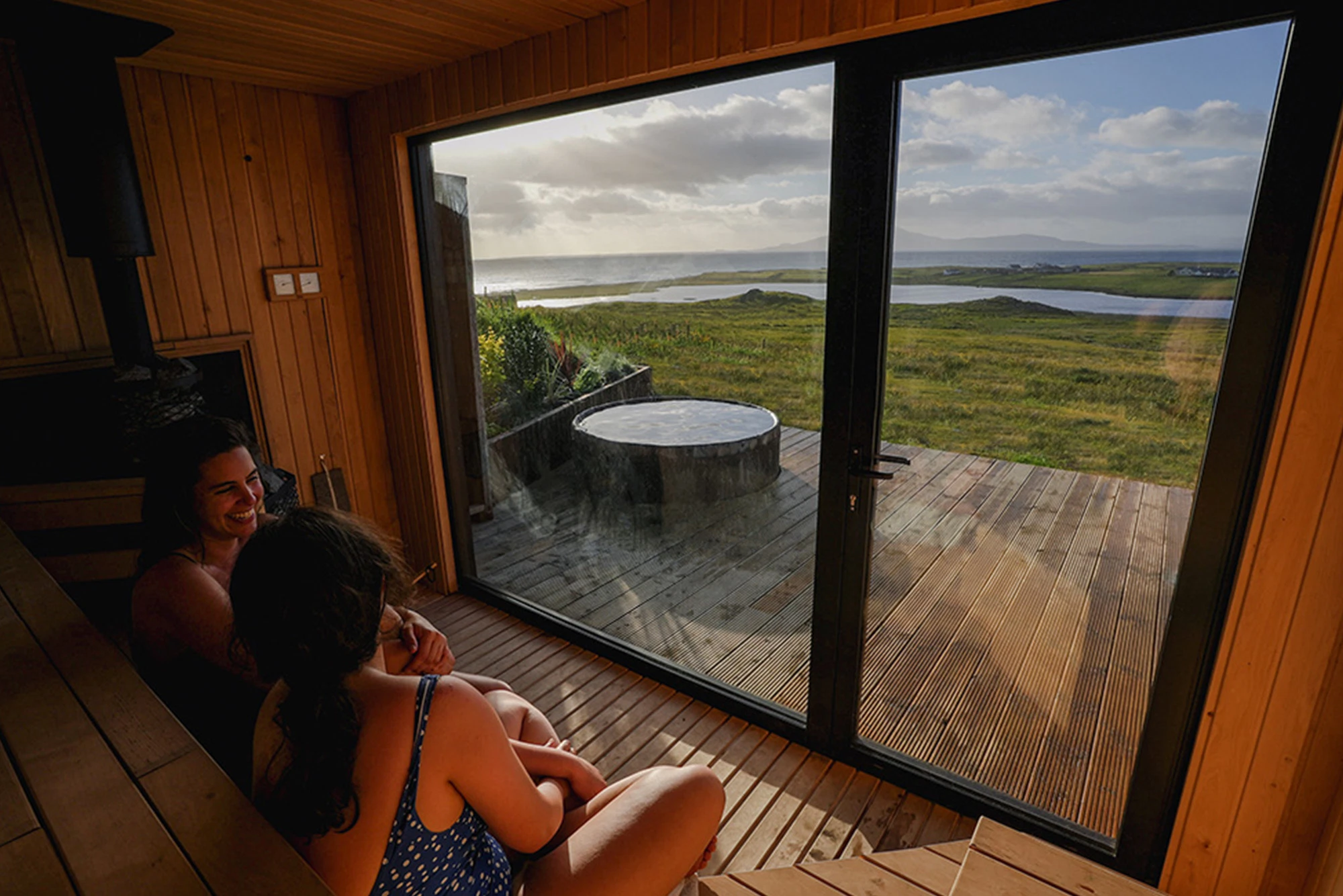 Two women in a wooden sauna enjoy coastal views through a glass door, overlooking a hot tub and scenic landscape.