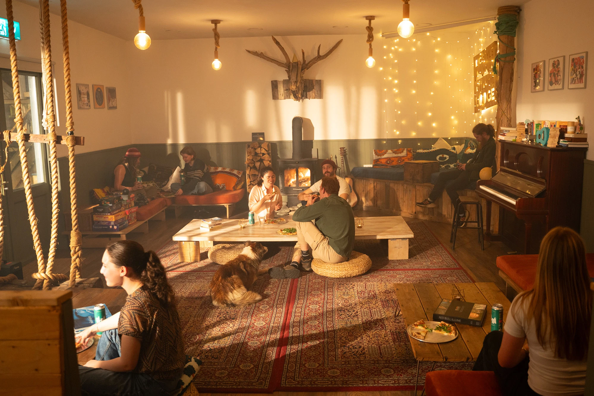 Cosy lounge with people socialising, eating pizza, and playing games near a wood stove and fairy lights.