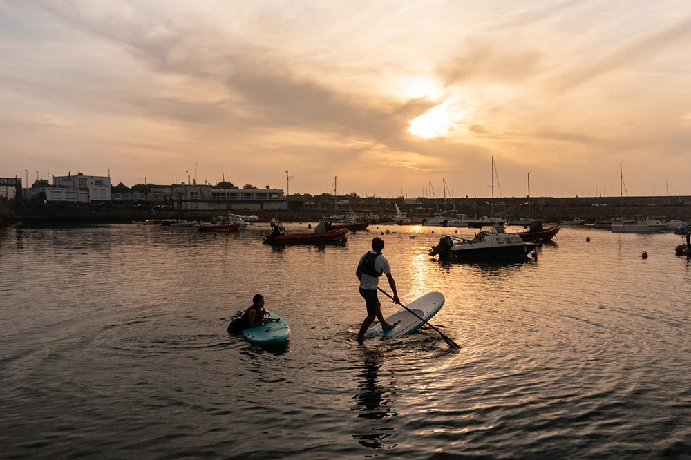 Sunset kayaking and paddleboarding in a tranquil harbour, boats moored under an orange sky.