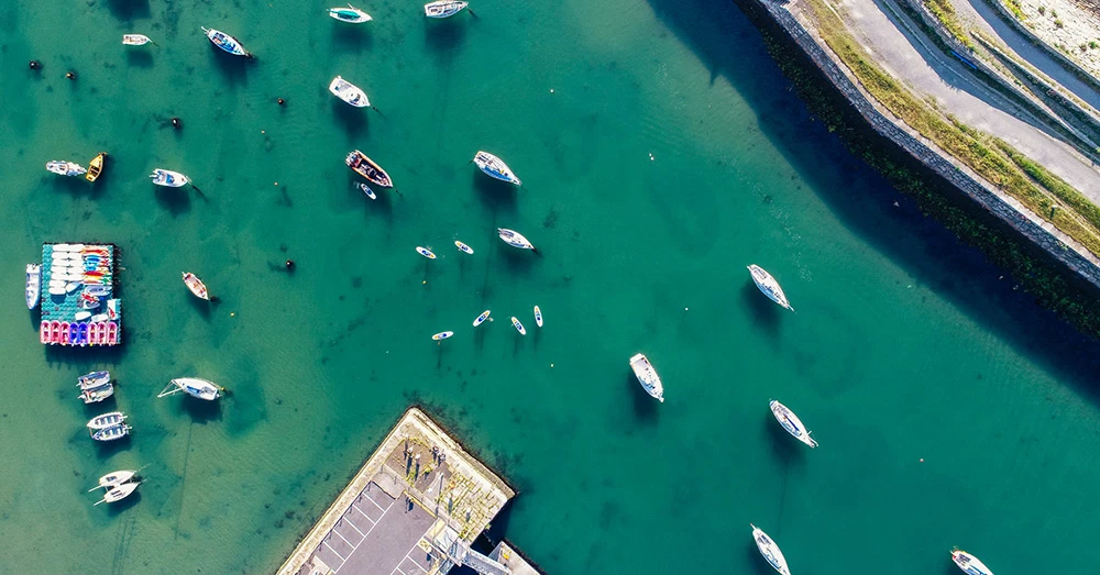 Aerial view of small boats moored on a tranquil, turquoise marina near a car park and stone road.
