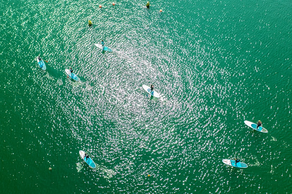 Aerial view of paddleboarders on a sunlit, emerald green sea.