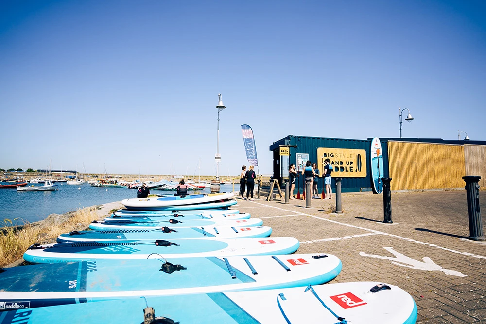 Stand up paddleboards lined up at a seaside rental shop with people chatting under clear blue skies.