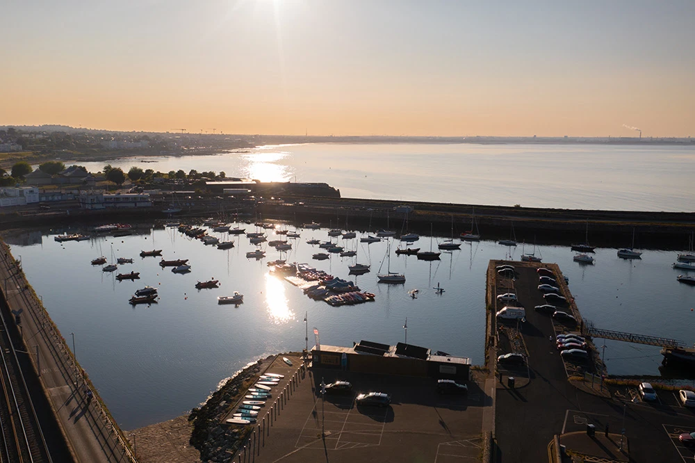 Aerial view of sunlit Dun Laoghaire Harbour with moored boats and coastal scenery at sunrise.