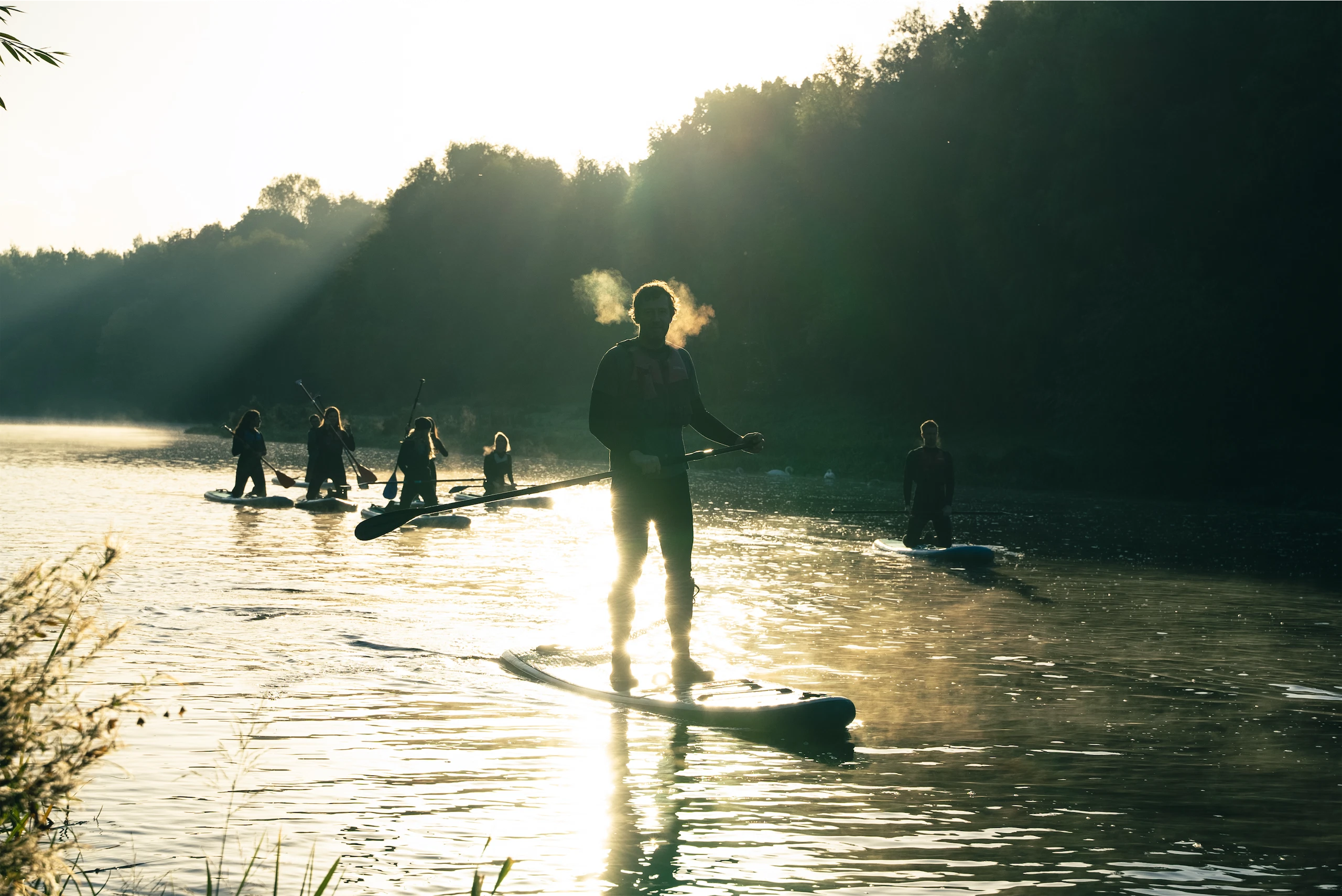 Silhouetted group paddleboarding on a serene river at sunrise, mist rising from the water.