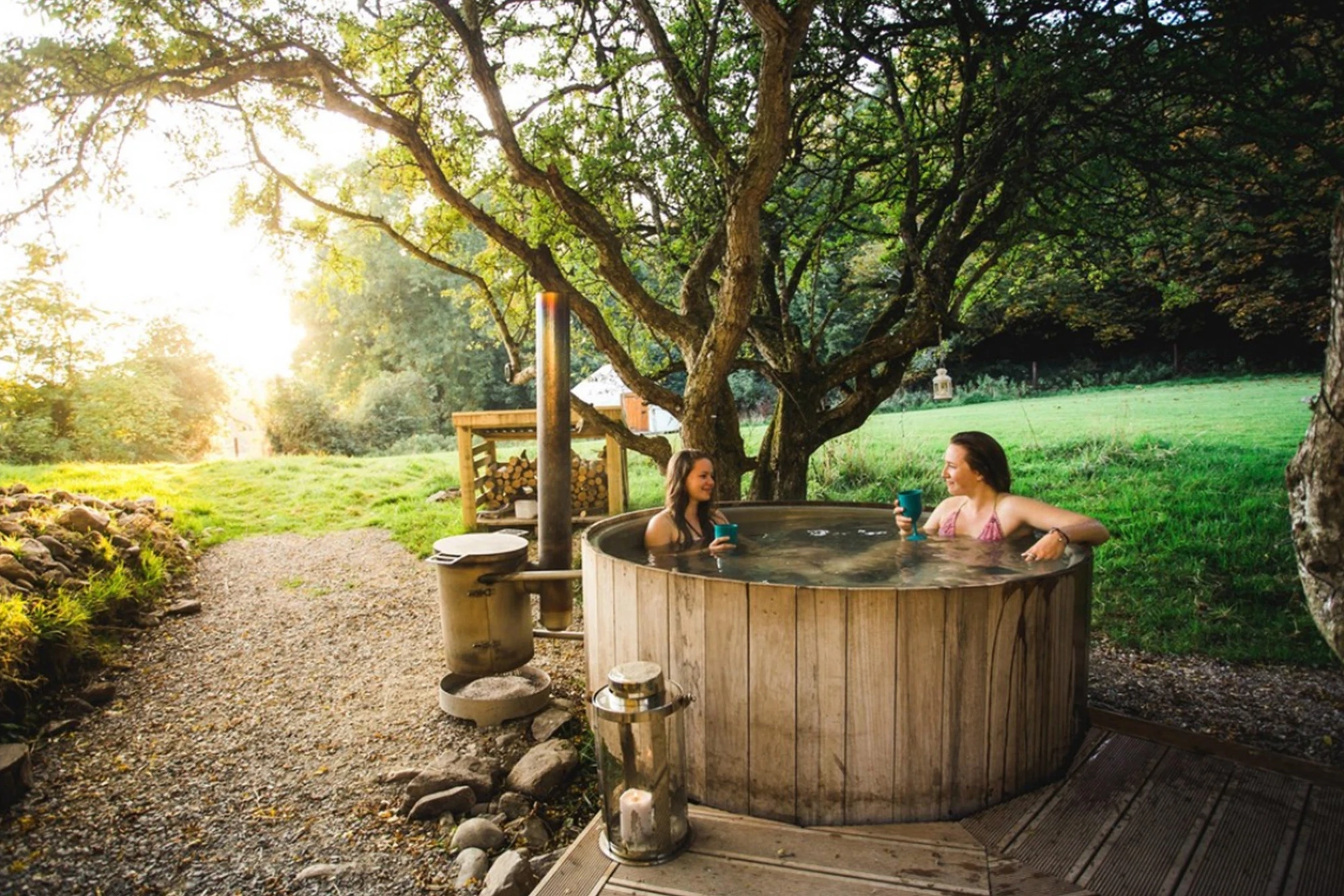 Two women enjoying a hot tub outdoors amid trees and soft evening light, holding drinks, in a tranquil rural setting.