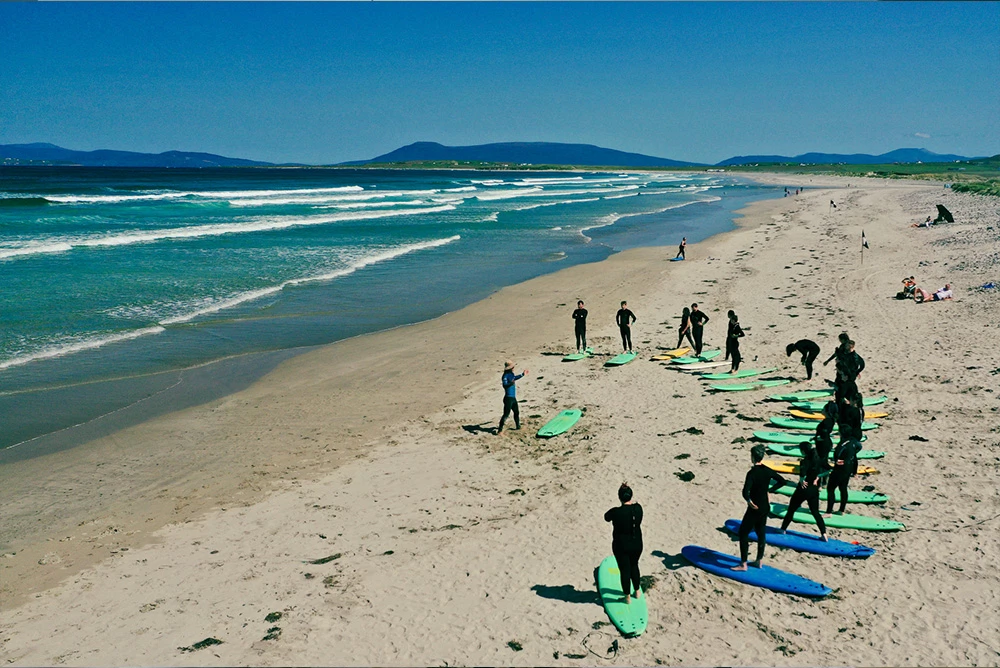 Surfers in wetsuits prepare on Donegal beach, Ireland. Blue skies, waves, and surfboards highlight coastal adventure.
