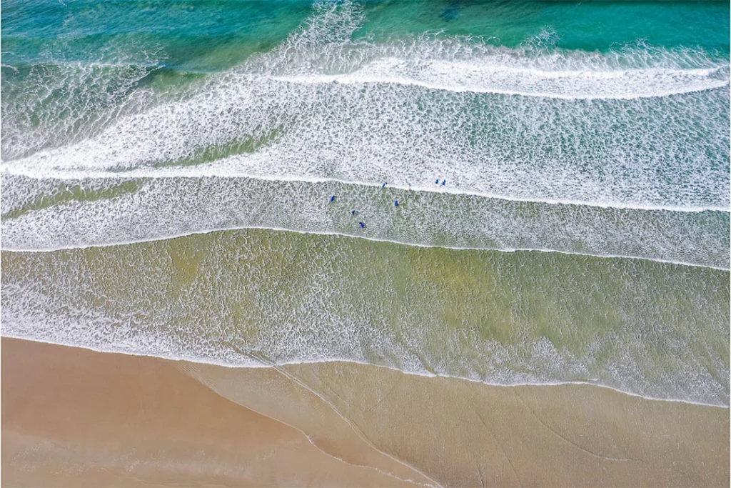 Aerial view of surfers in blue suits riding gentle waves at Inchydoney Beach, Ireland. Sandy shoreline visible.