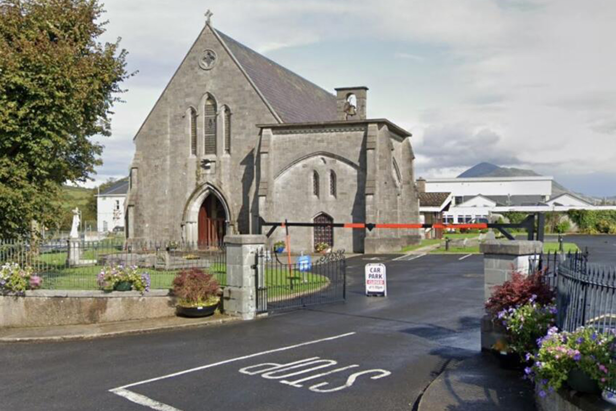 Historic stone church with Gothic architecture, fenced garden, and Car Park sign, set against a mountainous backdrop.