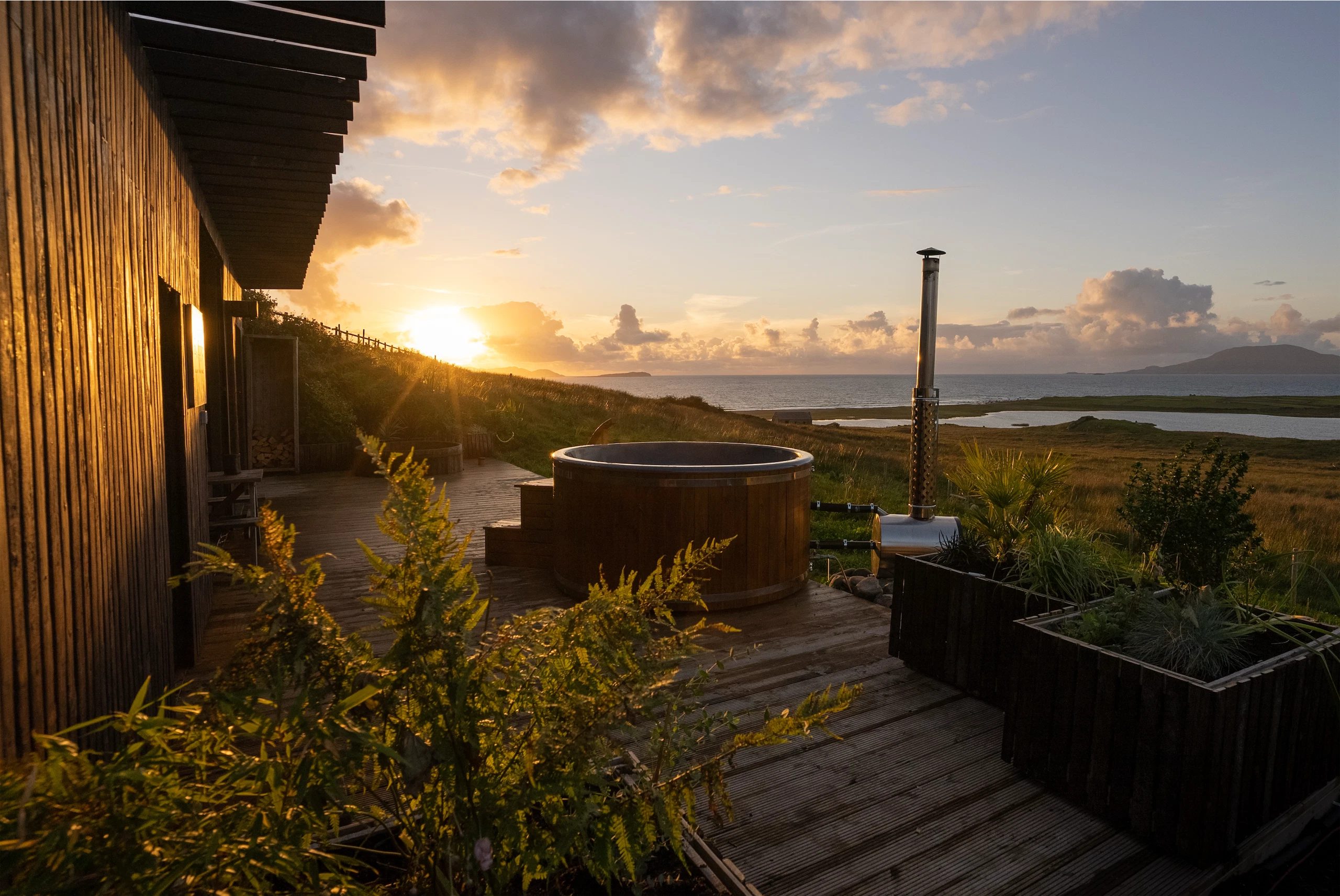 Sunset over coastal landscape with hot tub and wooden cabin, overlooking the sea.