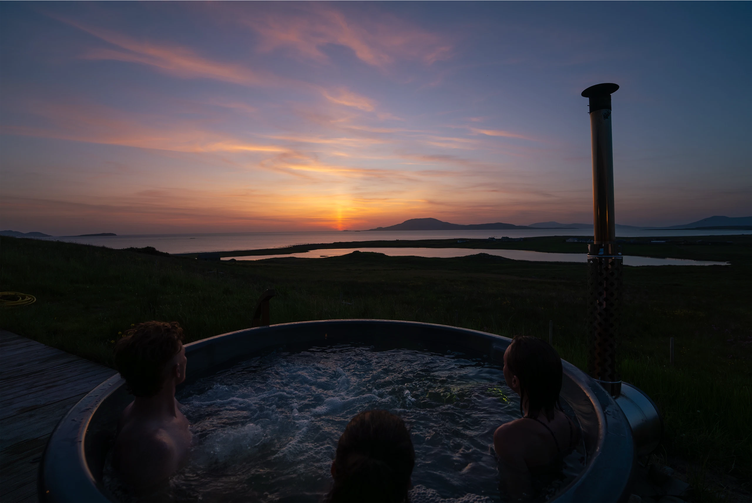 People enjoying a hot tub with a stunning sunset view over Irish coastline and hills.