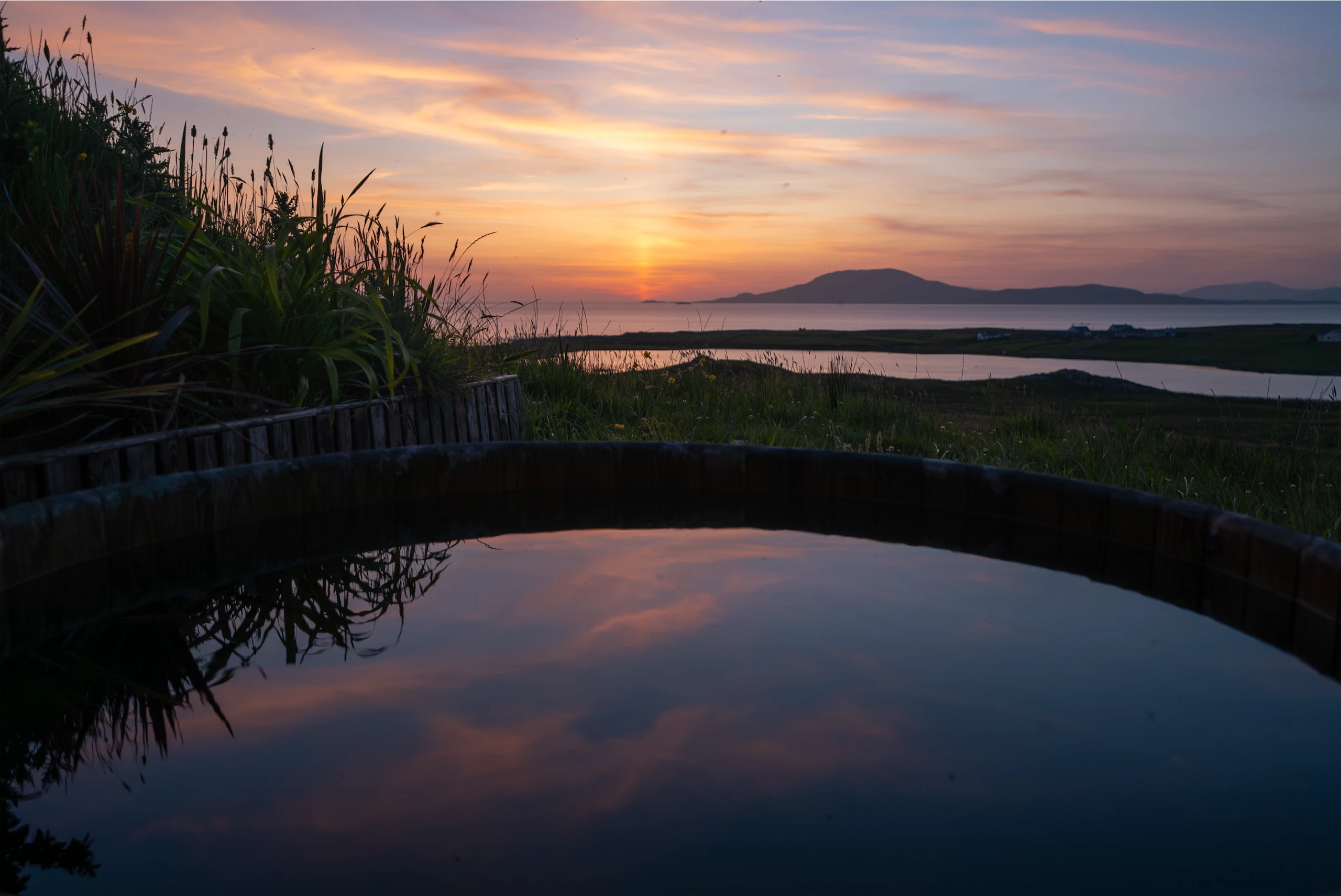 Sunset over calm sea with silhouetted hills; sky reflects in water.