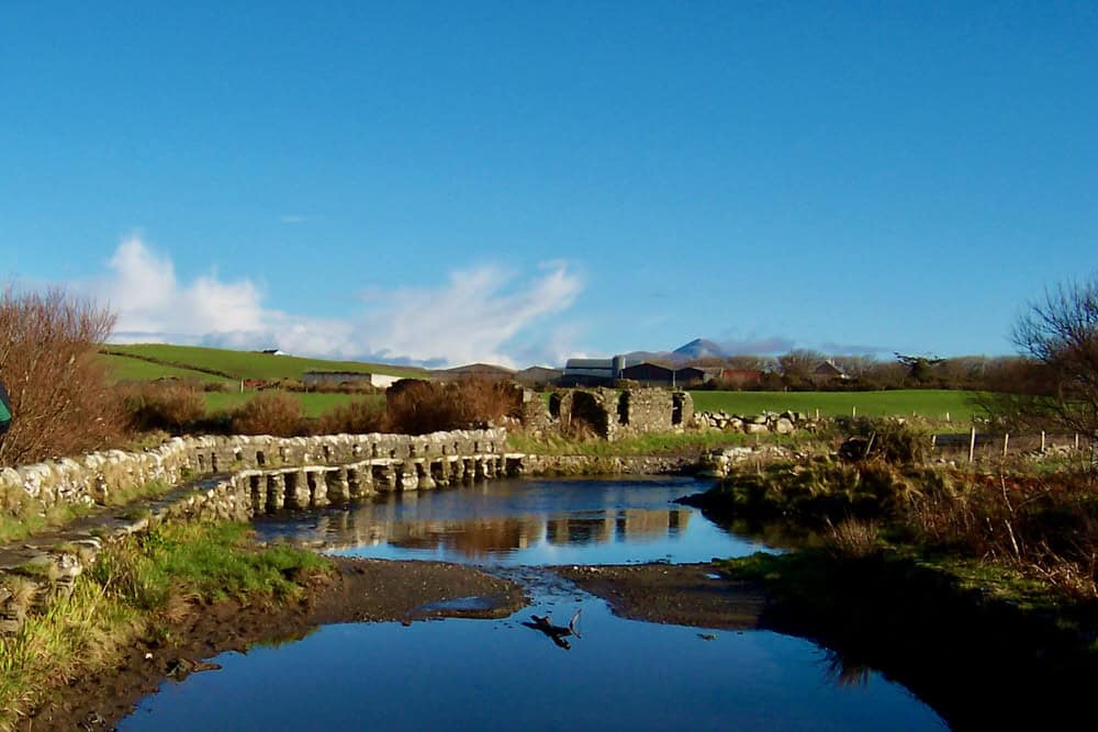 Stone bridge over a calm stream in Irish countryside with hills and blue sky in the background.