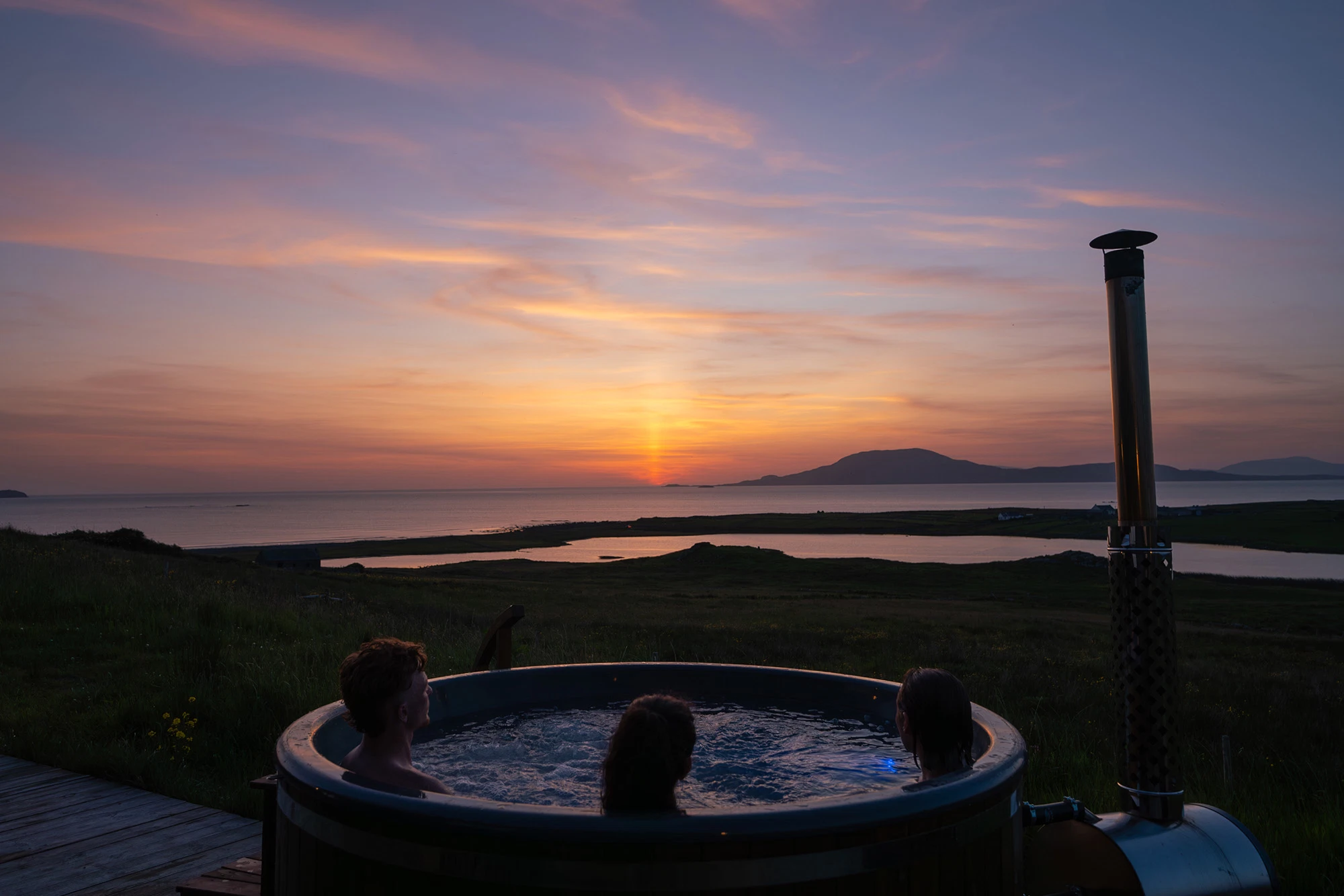 People relaxing in a hot tub overlooking a scenic coastal sunset.