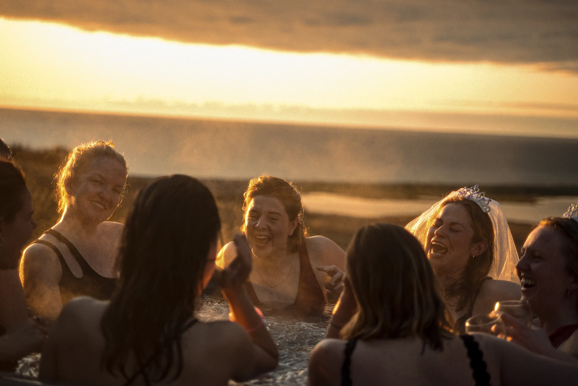 Hen party laughing in a hot tub during sunset, with tiaras and drinks, overlooking a scenic ocean view.