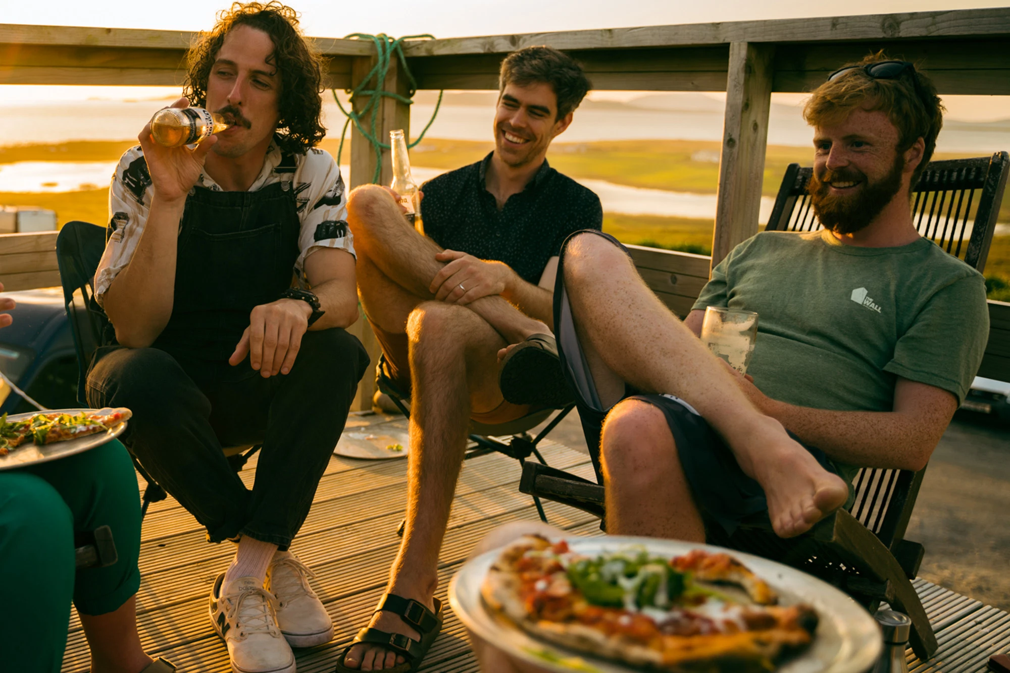 Three friends relaxing on a sunny veranda, enjoying drinks with a scenic coastal view.