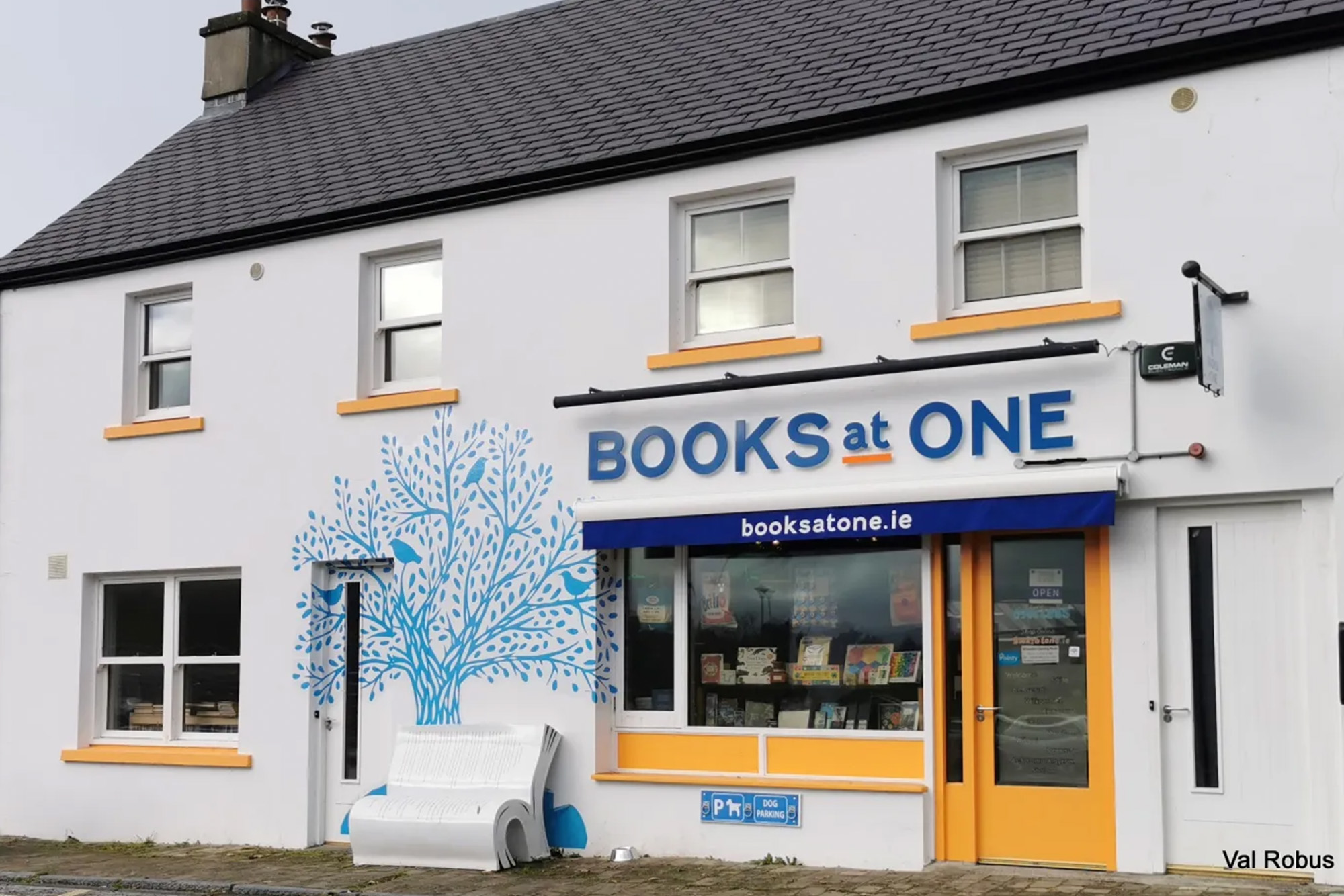 White bookshop with a blue tree mural and a unique open book bench. Sign reads Books at One.