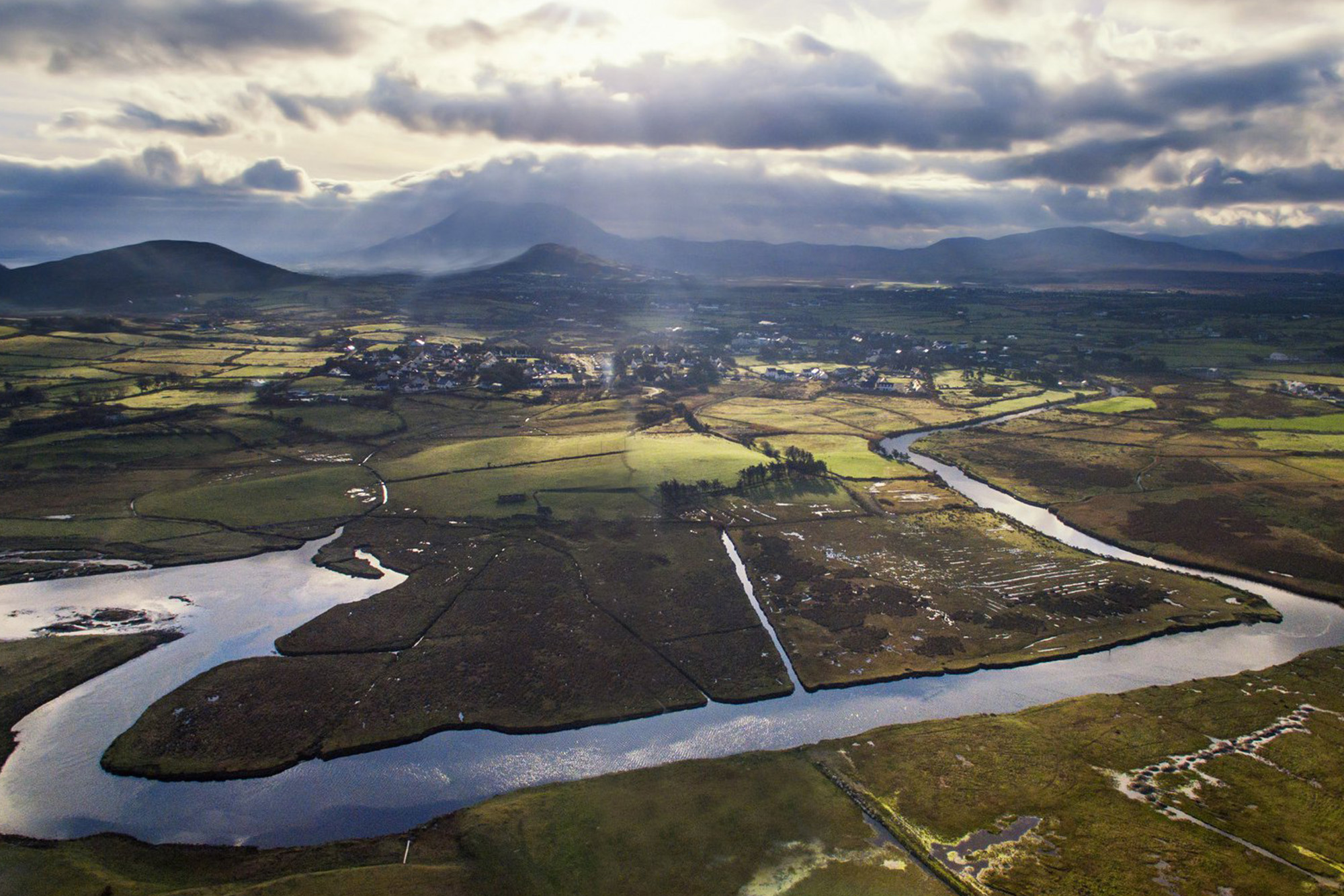 Aerial view of scenic Irish landscape with winding river, fields, and distant hills under a cloudy sky.