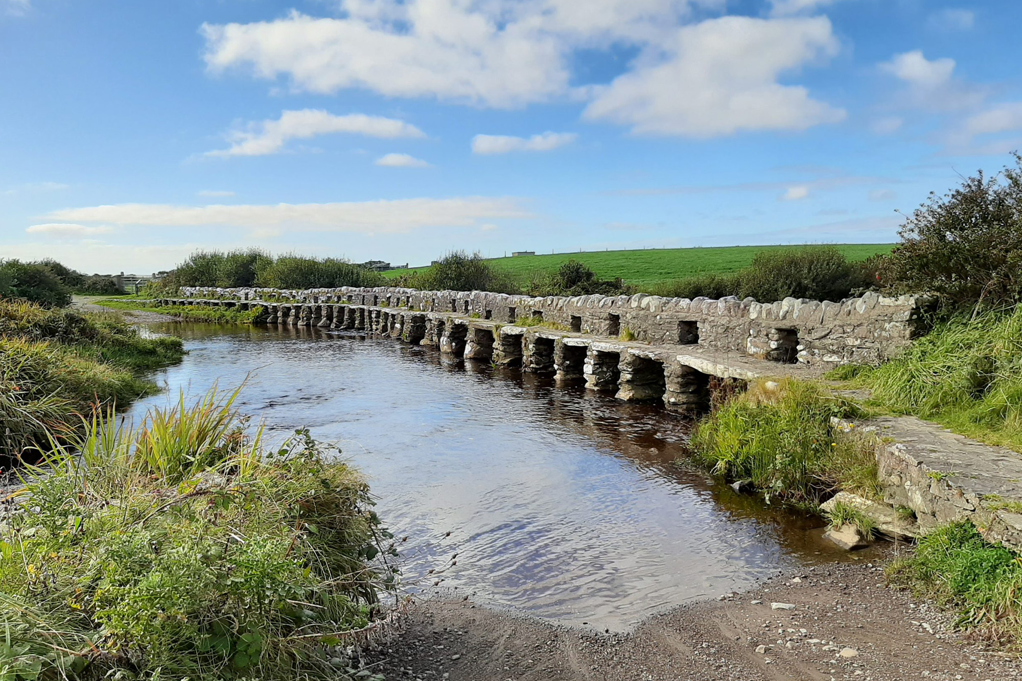 Historic clapper bridge over a tranquil river, with lush green fields and a bright blue sky in the background.