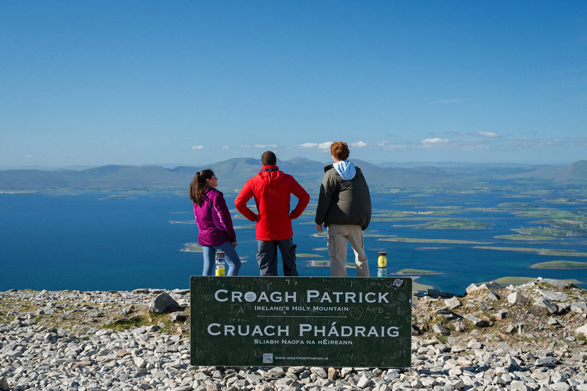 Three hikers stand at Croagh Patrick summit, overlooking Clew Bay's islands and distant mountains on a clear day.