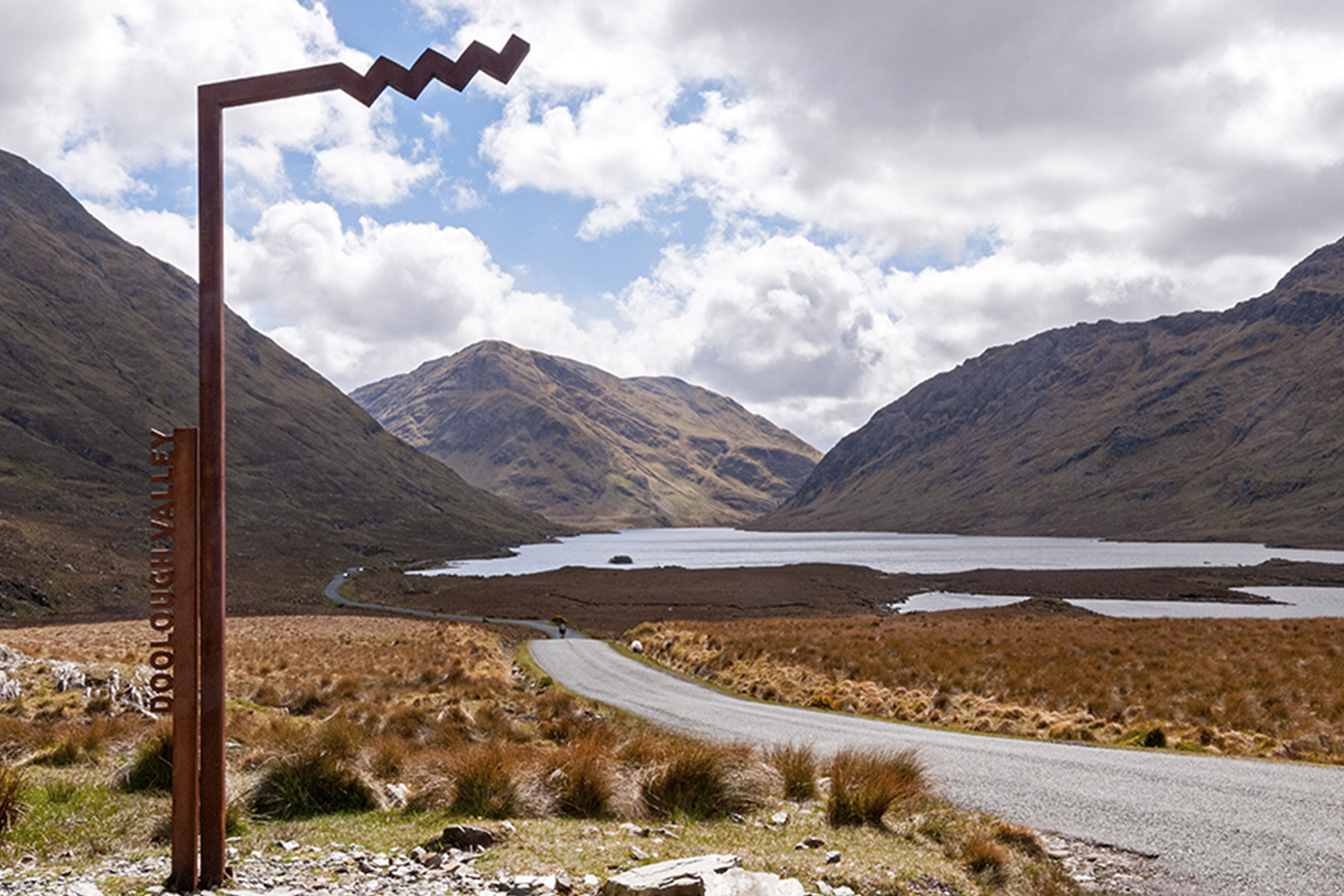 Doolough Valley scenic view with mountains, lake, and distinctive metal sign under a cloudy sky.
