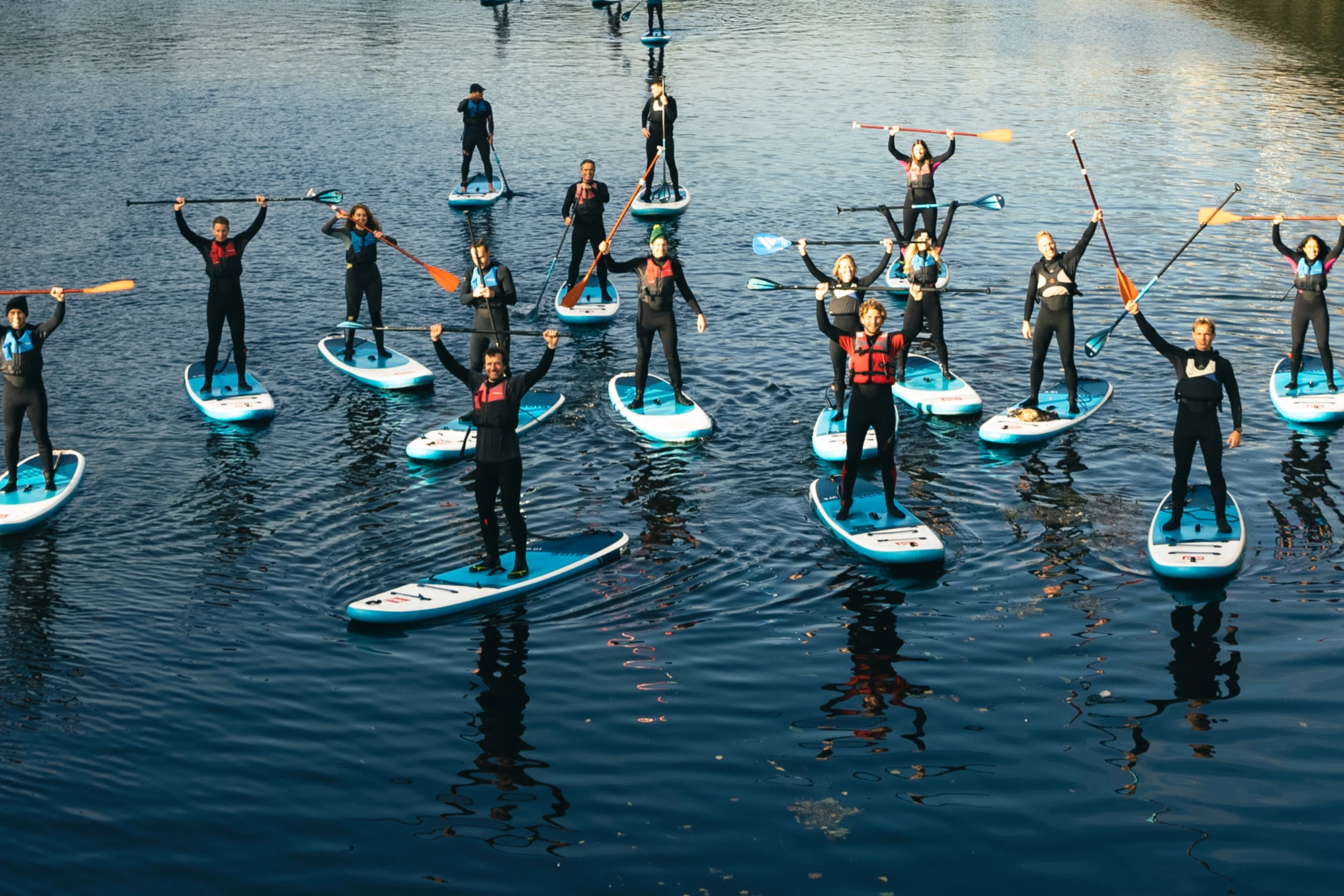 Group paddleboarding on calm water, participants in wetsuits holding paddles overhead.