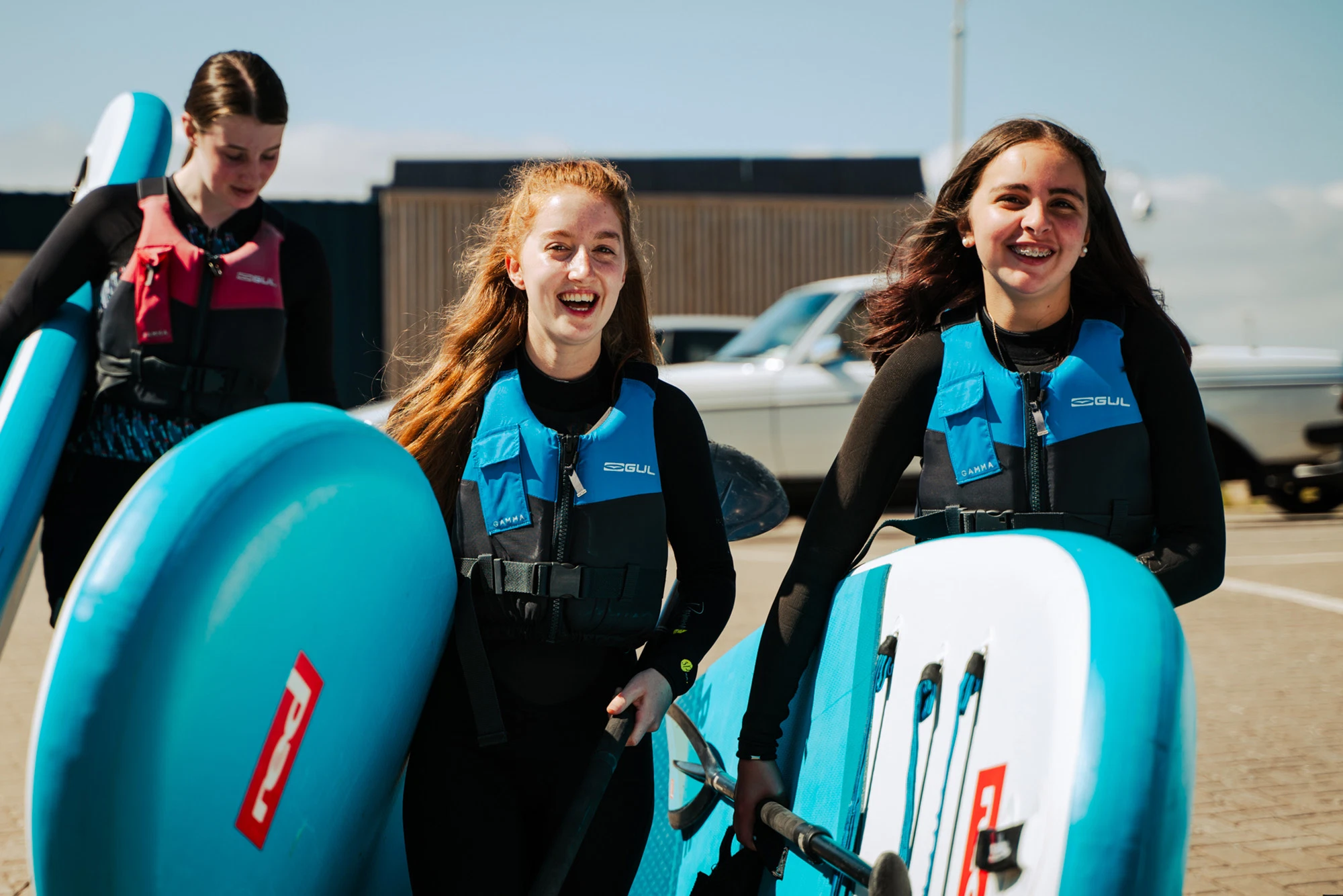 Three women in wetsuits and life jackets carry surfboards, smiling under a clear sky.