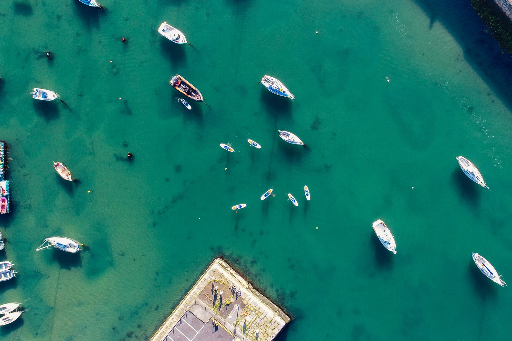 Aerial view of sailboats moored in turquoise harbour water beside a stone jetty, creating a serene coastal scene.