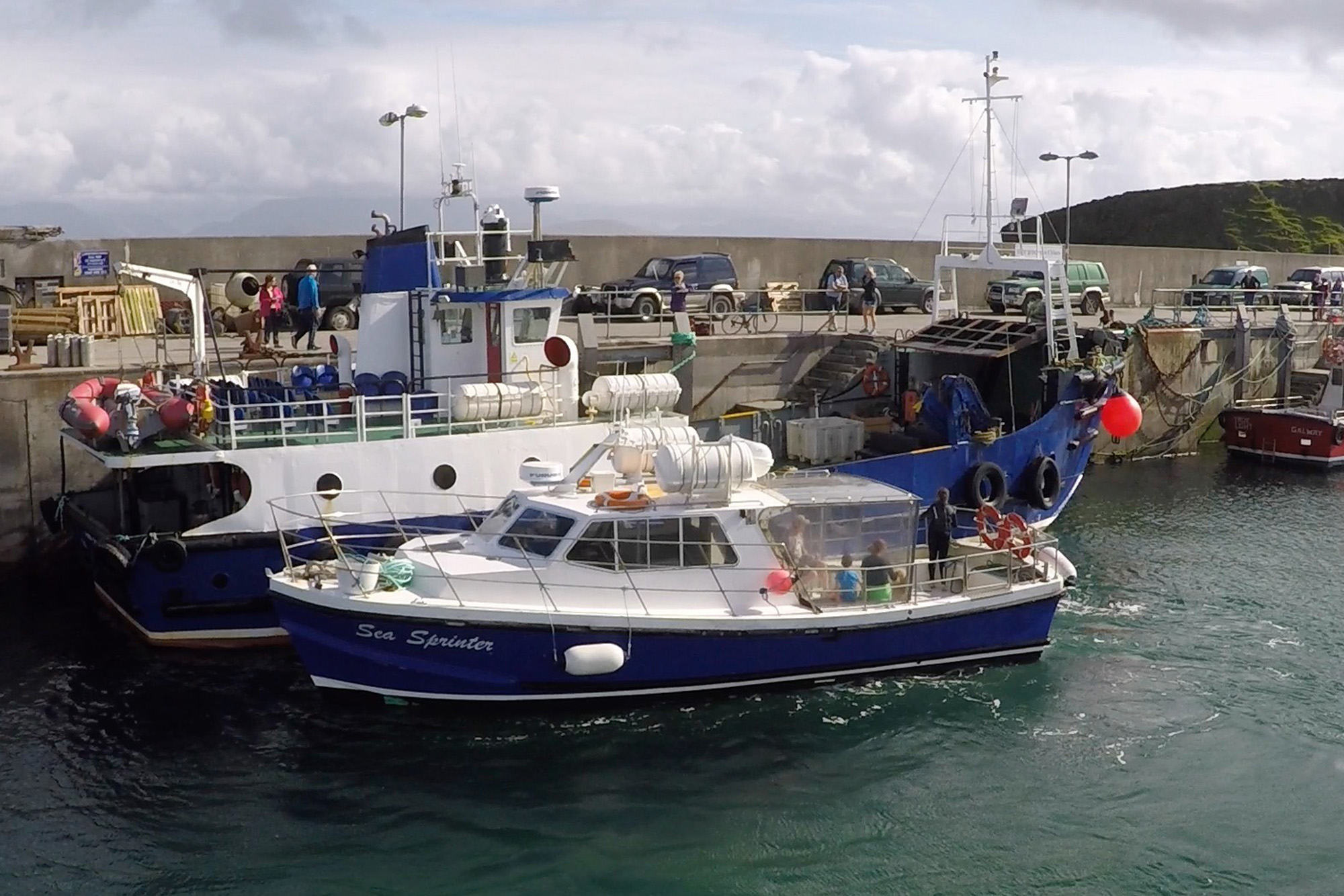 Boats docked at Irish harbour; people on quay with cars. Overcast sky, fishing gear visible.
