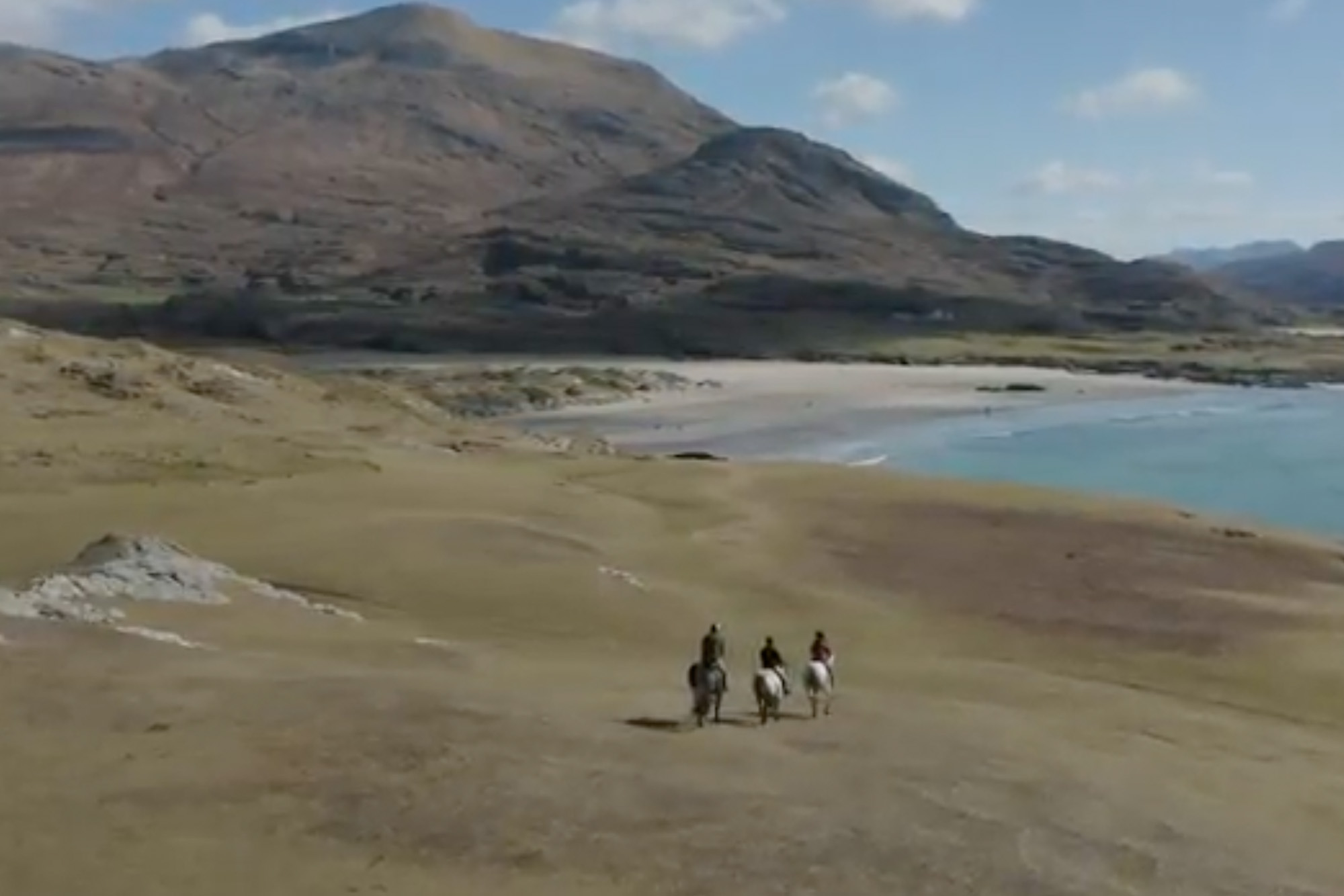 Three horse riders traverse scenic Irish coastline with mountains and beach in the background.