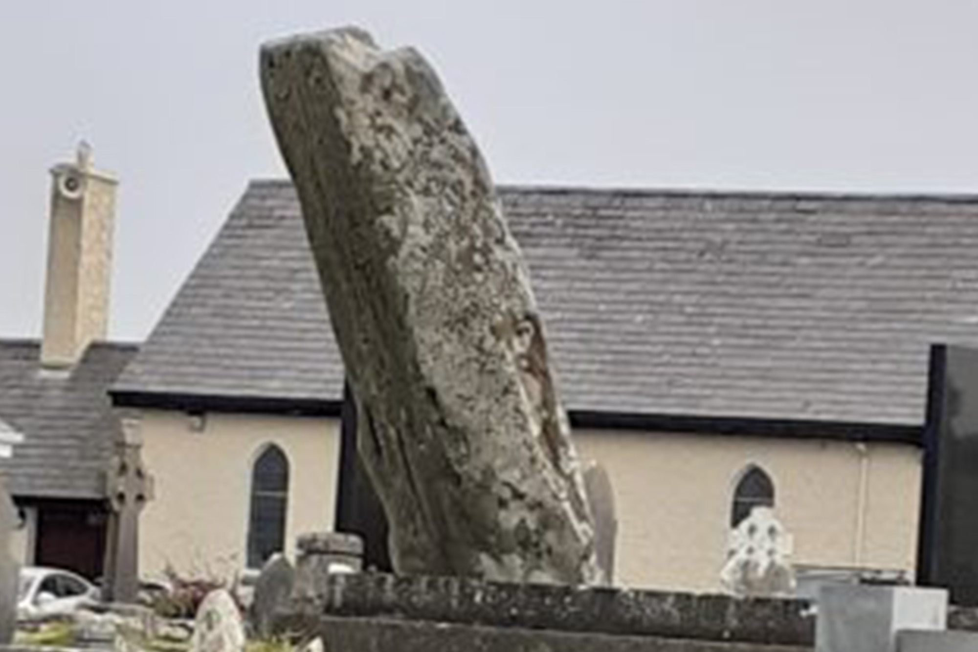 Leaning ancient gravestone in churchyard with stone church and chimney in background.