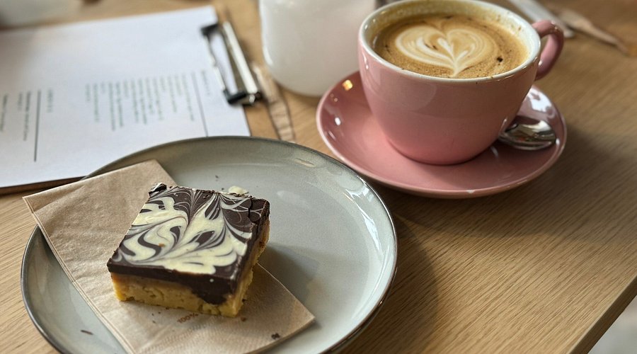 Coffee in pink cup with latte art, beside a marble chocolate slice on a plate, on a wooden café table.