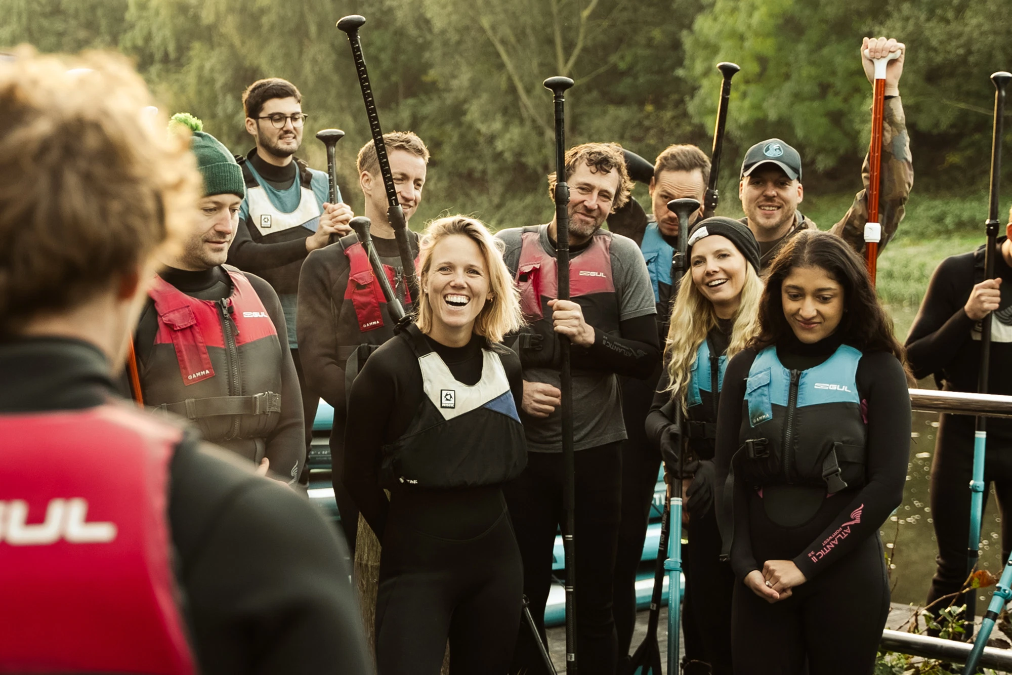 Group of smiling paddleboarders in wetsuits and life vests, holding paddles by a scenic river.