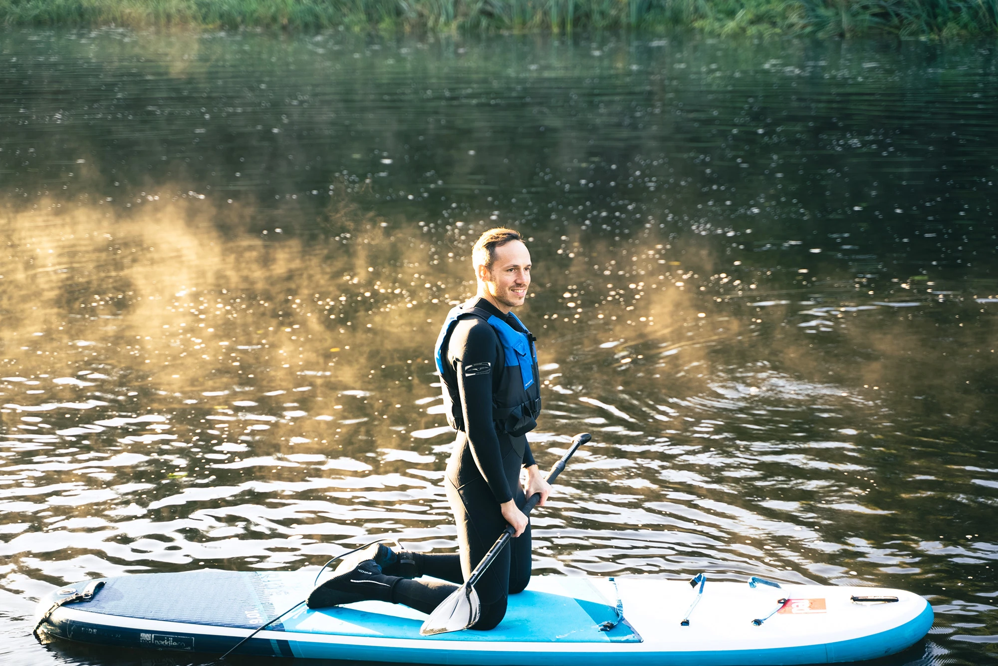 Man kneeling on paddleboard in wetsuit on calm lake, holding paddle with mist in background.