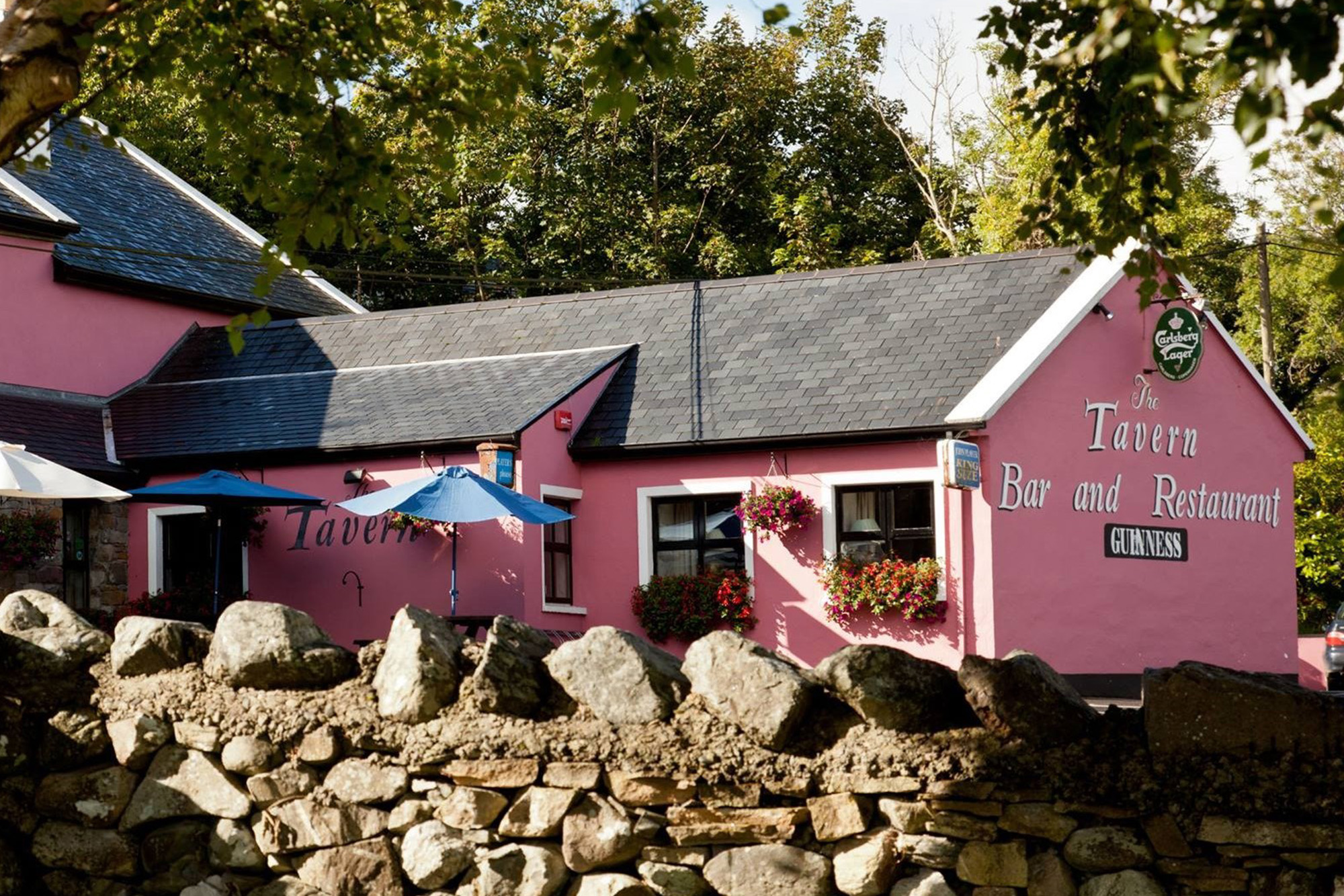 Pink tavern with patio umbrellas and hanging flowers, set behind a stone wall amidst lush trees.