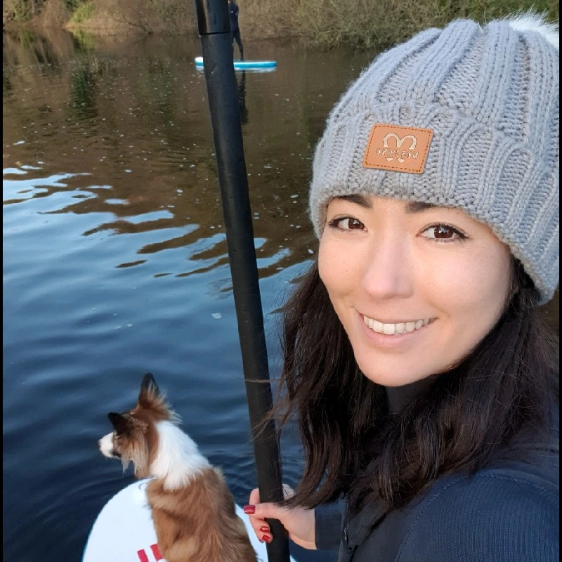 Woman paddleboarding with dog on calm river, wearing grey knit hat, smiling in winter setting.